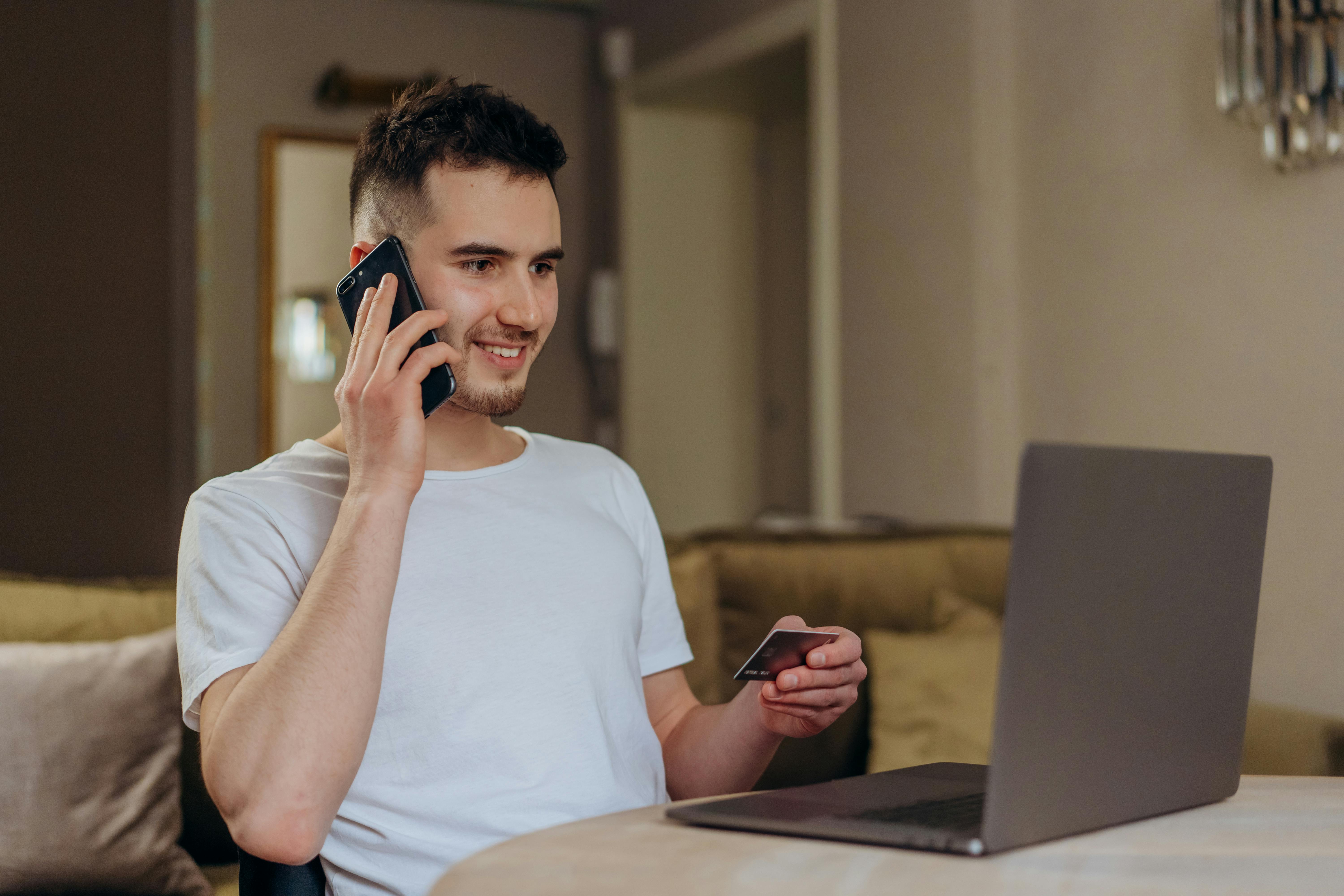 young person sitting on a sofa talking on a mobile phone on one hand and holding a credit card in the other, while looking at a laptop
