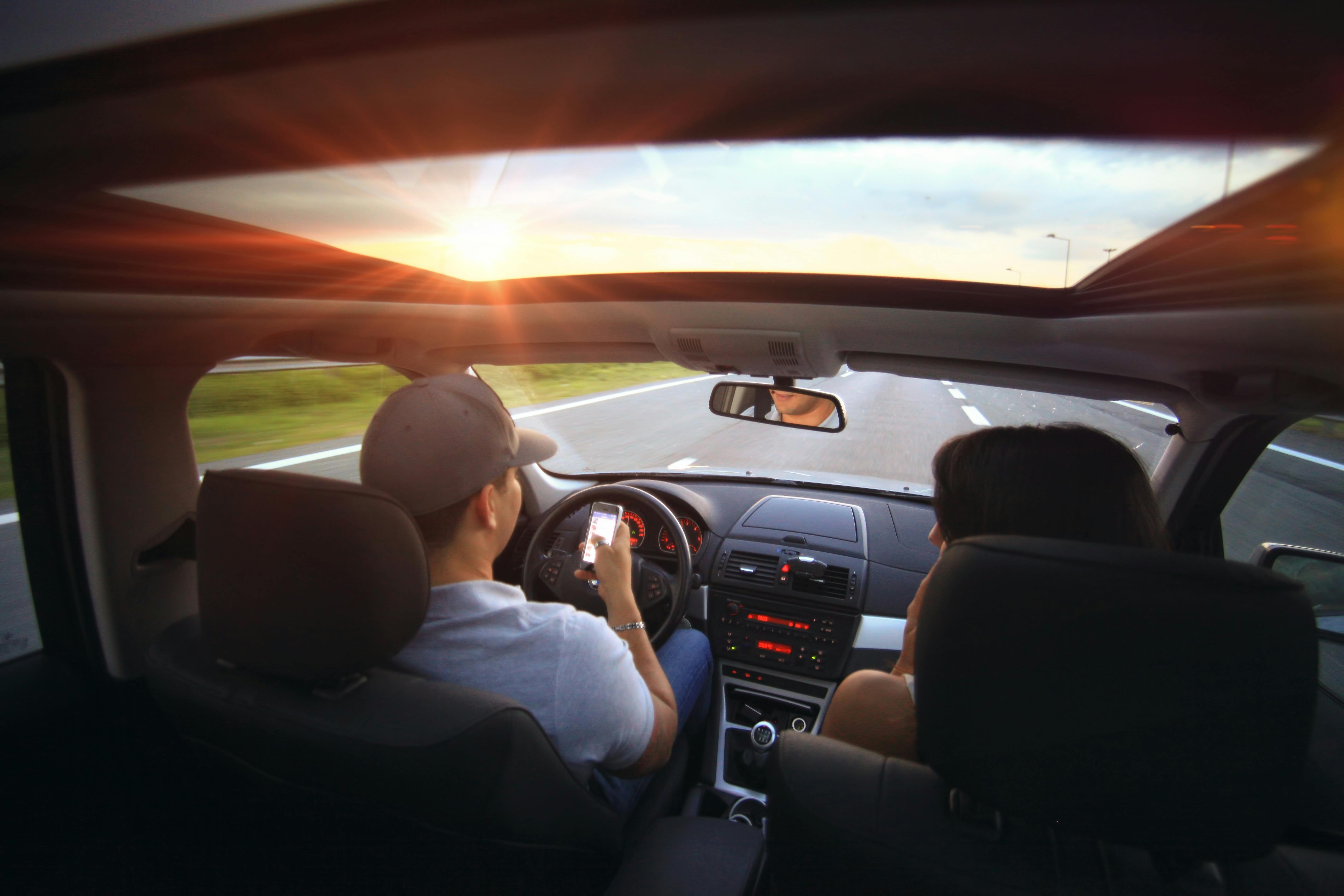 person driving while looking at a mobile phone, we can see him smiling in the rearview mirror, while his woman passenger is turned toward him, not the safest but at least they``re in the country side during a sun set