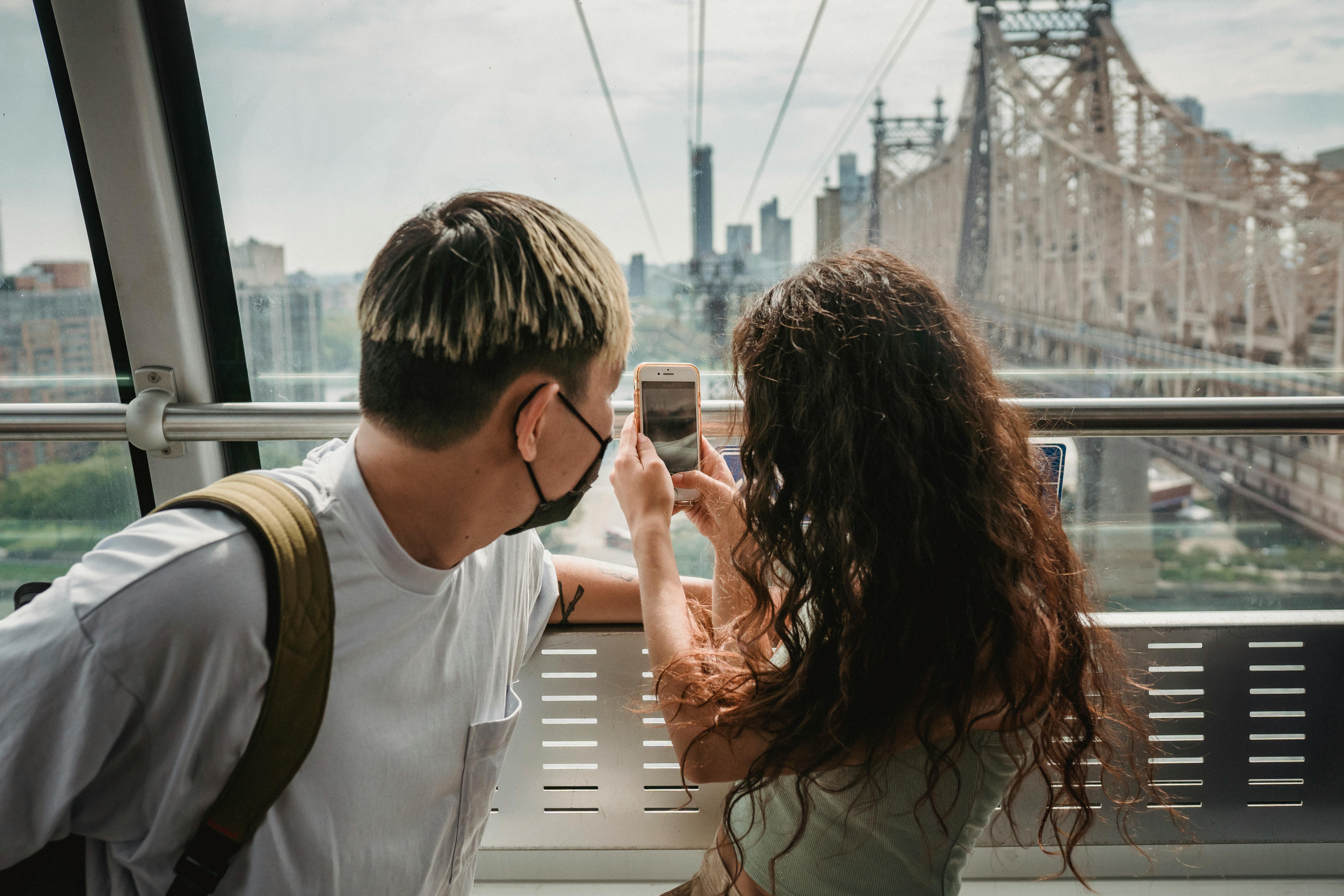 two people standing on a bridge, taking a picture of the city, while on their mobile phone