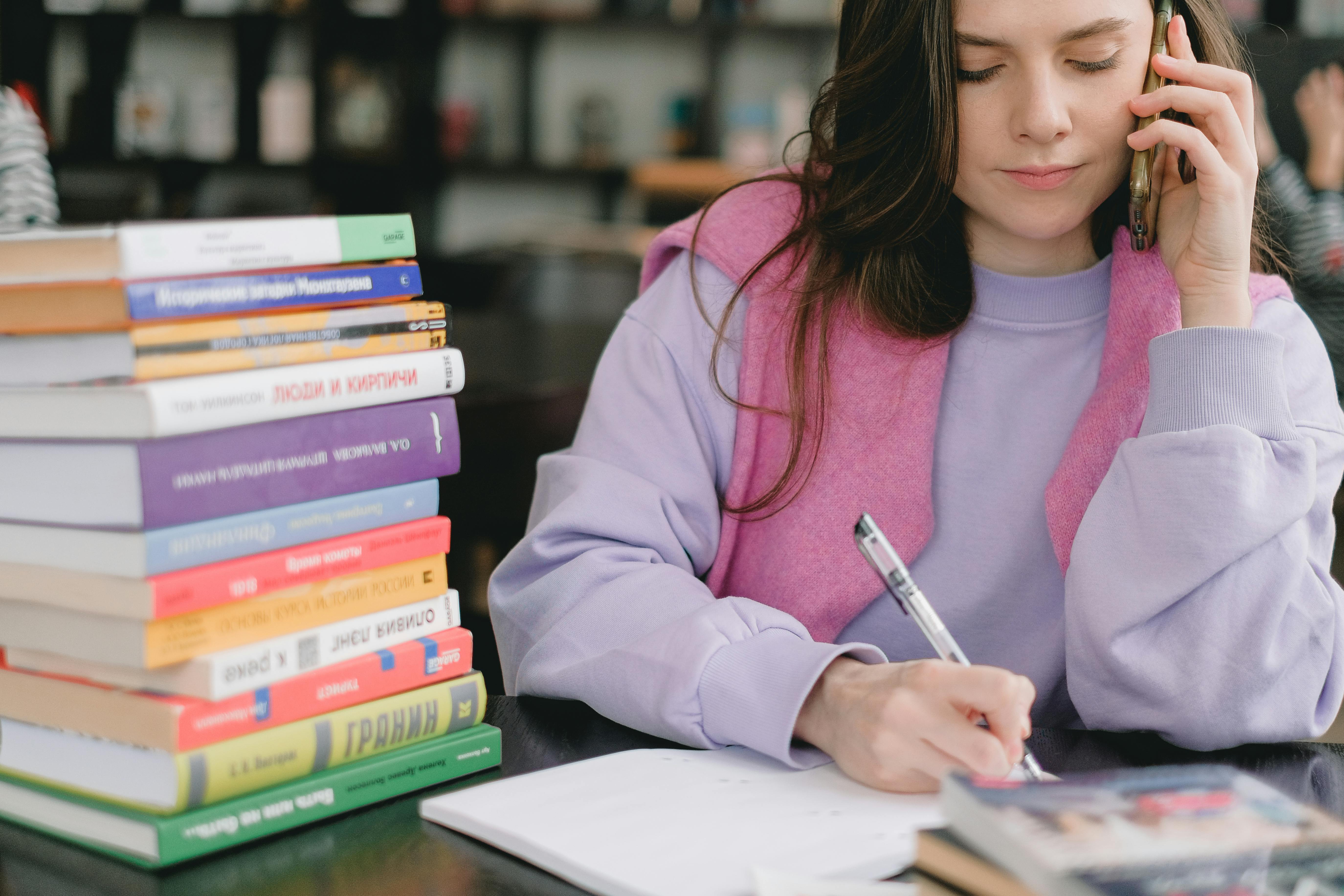 person on mobile phone taking notes with stack of books on desk
