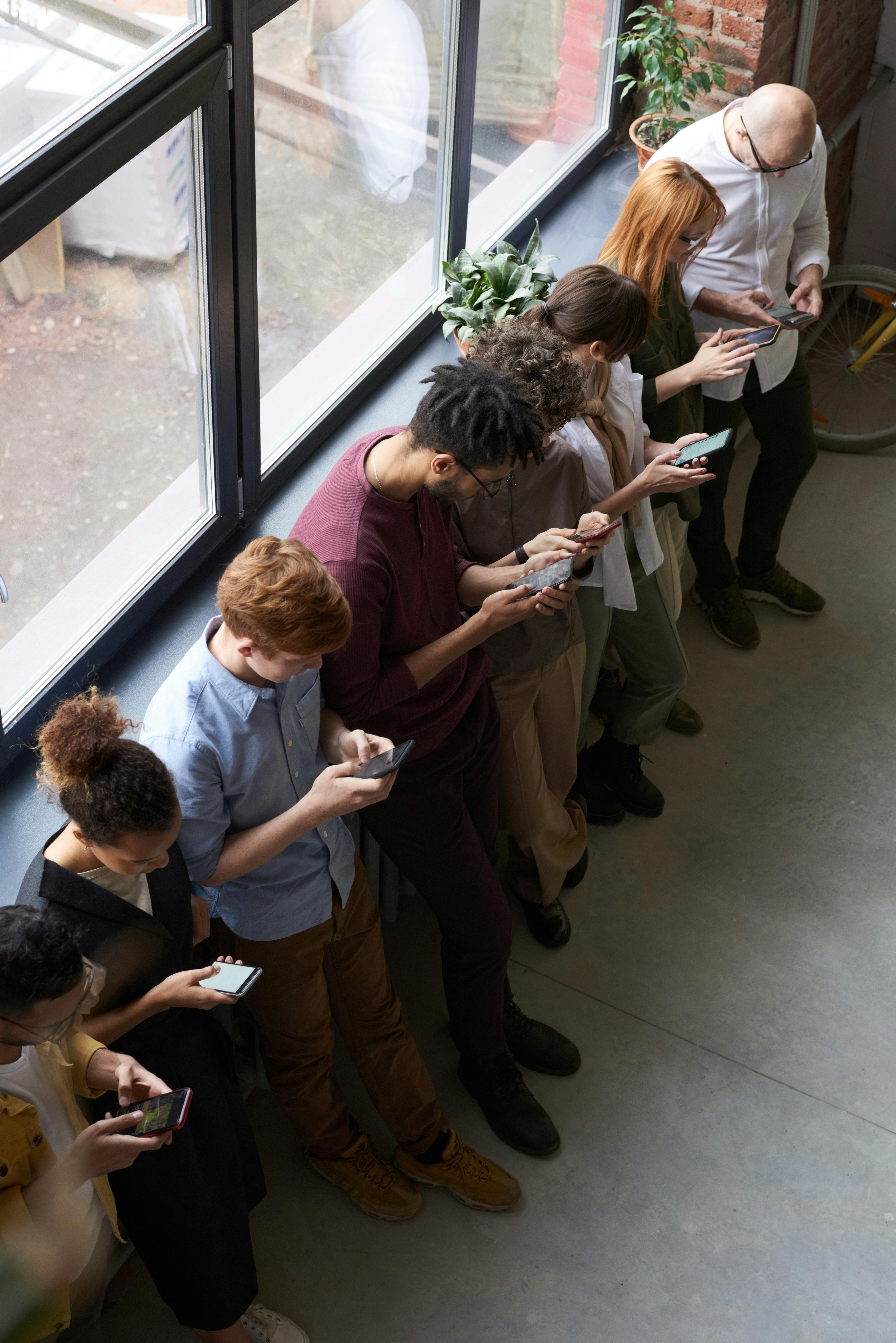 looking down on a crowd of people leaning against a wall, all of them looking at their mobile phones